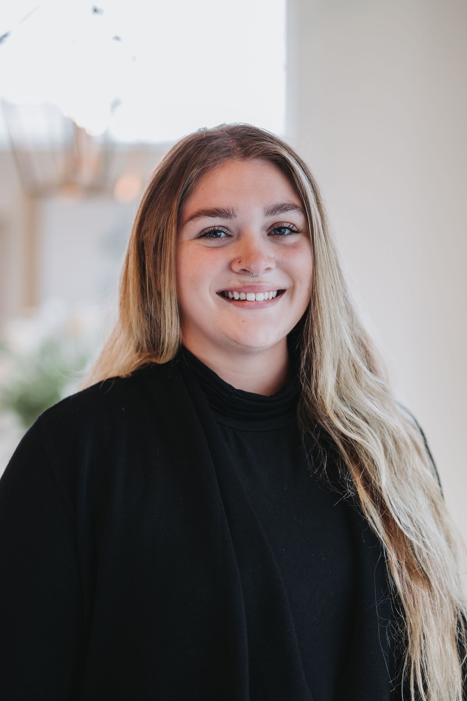 Smiling young woman with long blonde hair wearing a black turtleneck and blazer in a bright indoor setting.