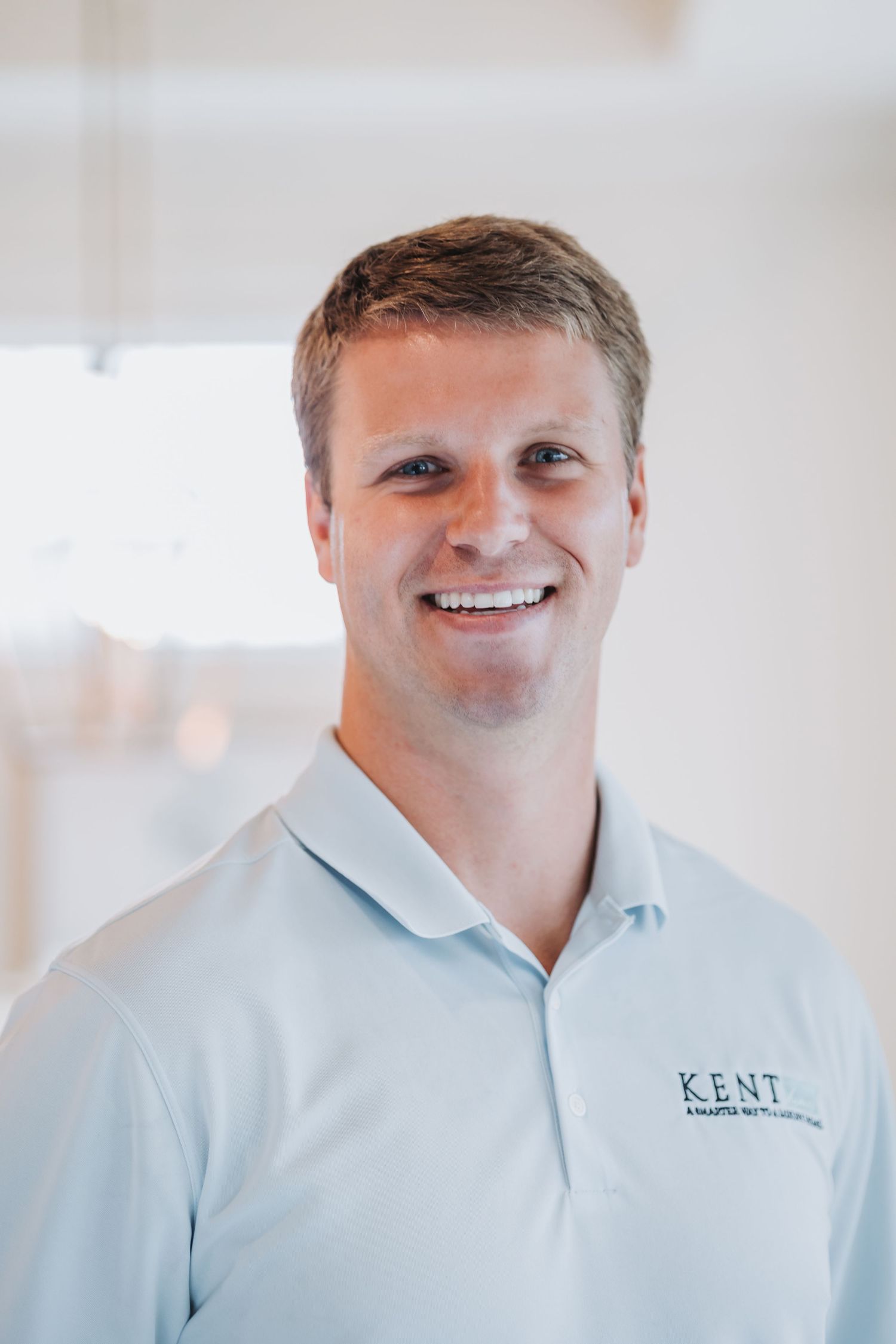 Smiling man in a light blue Kent polo shirt poses for a professional headshot in a bright office setting.