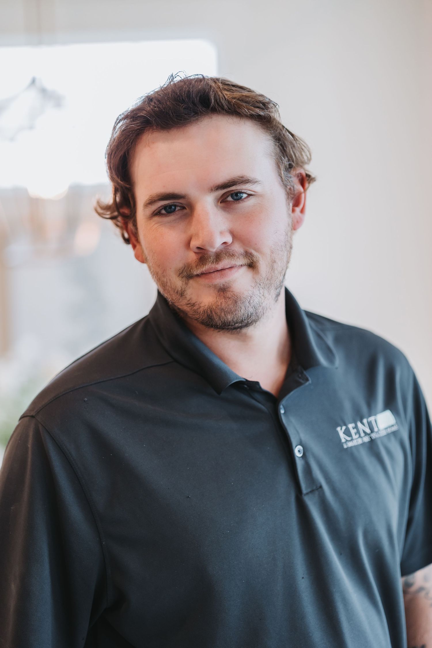 Young man with beard smiling in dark gray Kent polo shirt indoors.