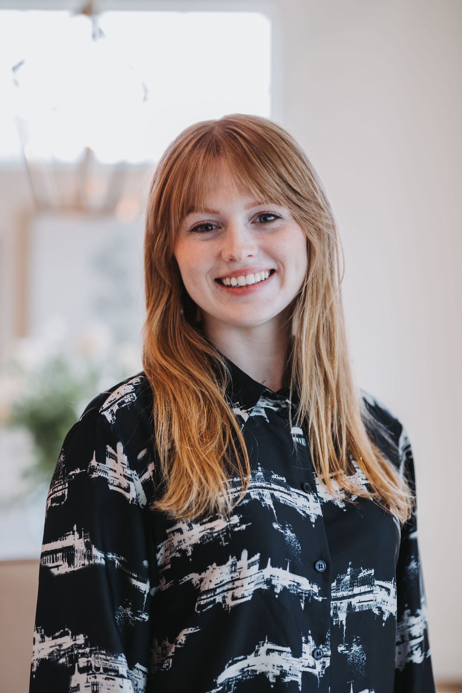 Smiling young woman with long red hair wearing a black patterned shirt, posing indoors in a bright room.