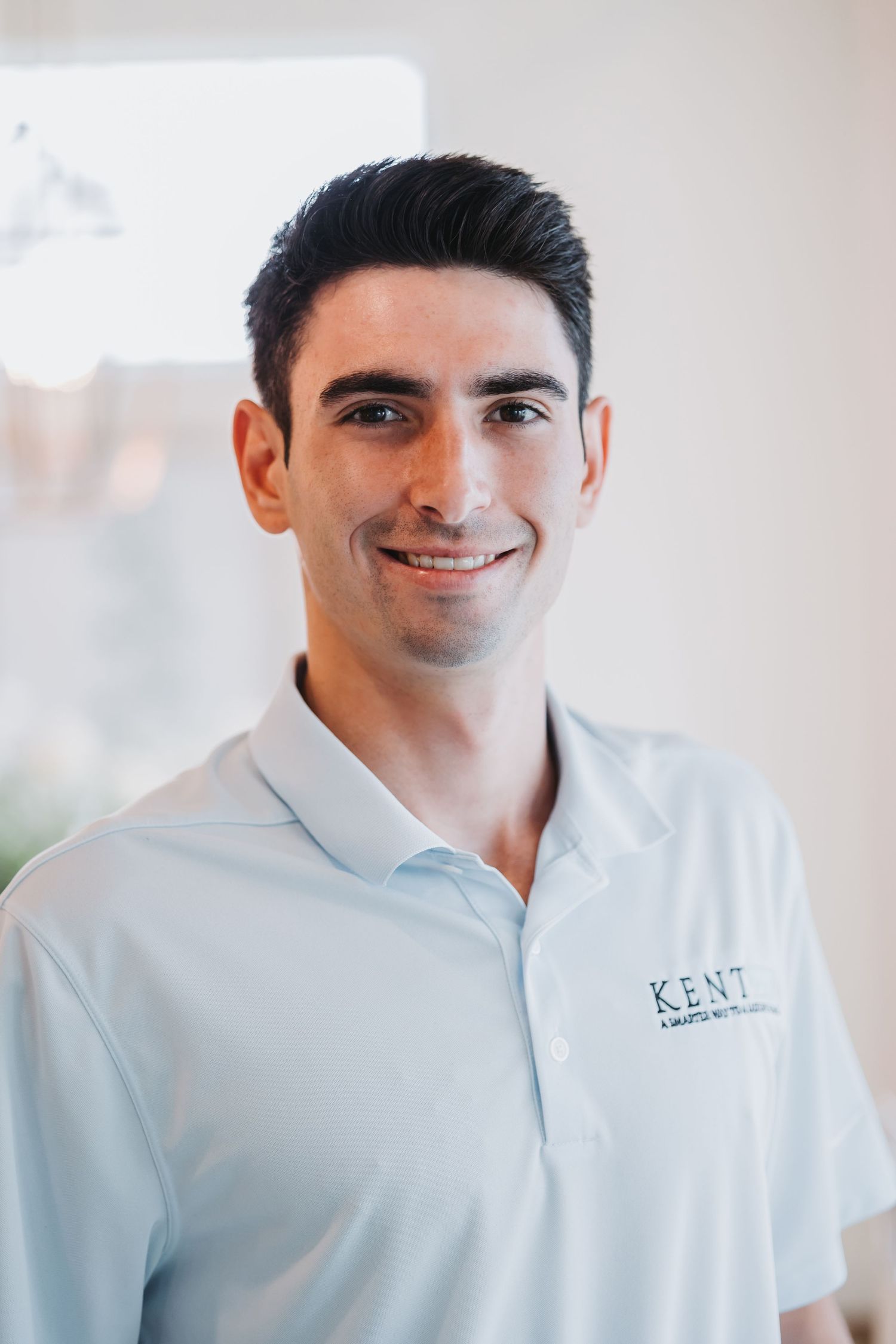 Smiling young man in a light blue Kent polo shirt posing indoors for a professional headshot photo.