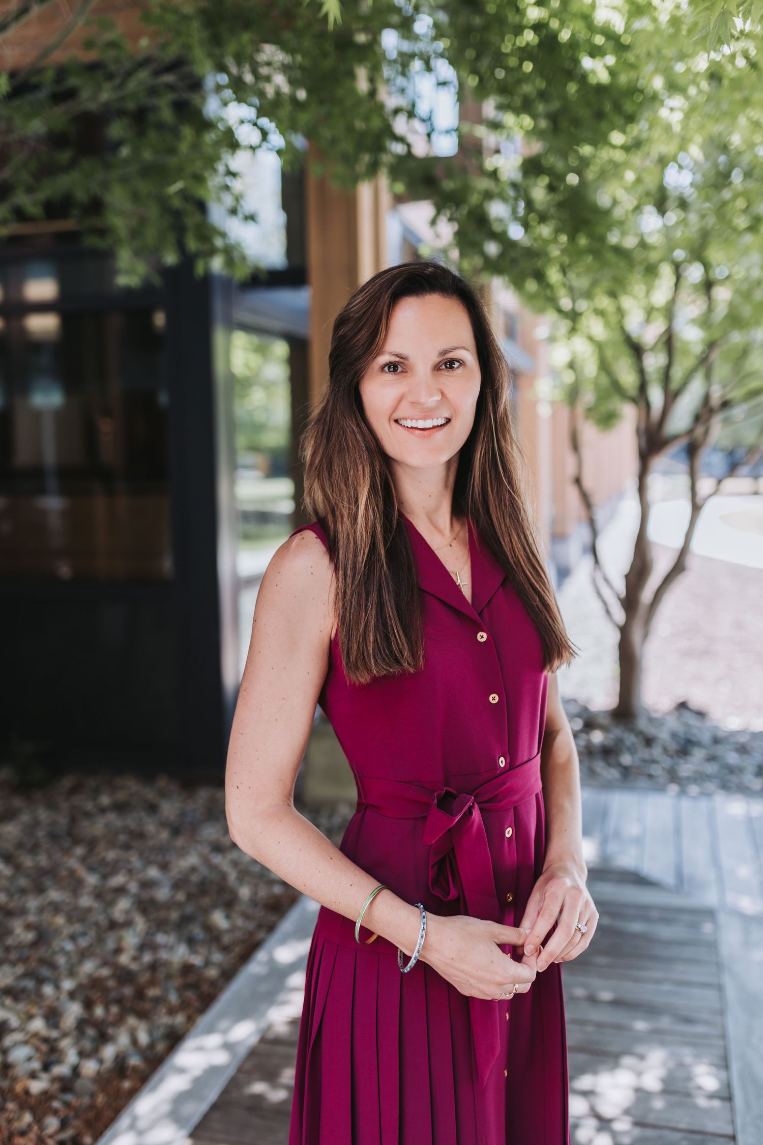 A smiling woman in a burgundy sleeveless dress stands outdoors near green trees and a modern building.