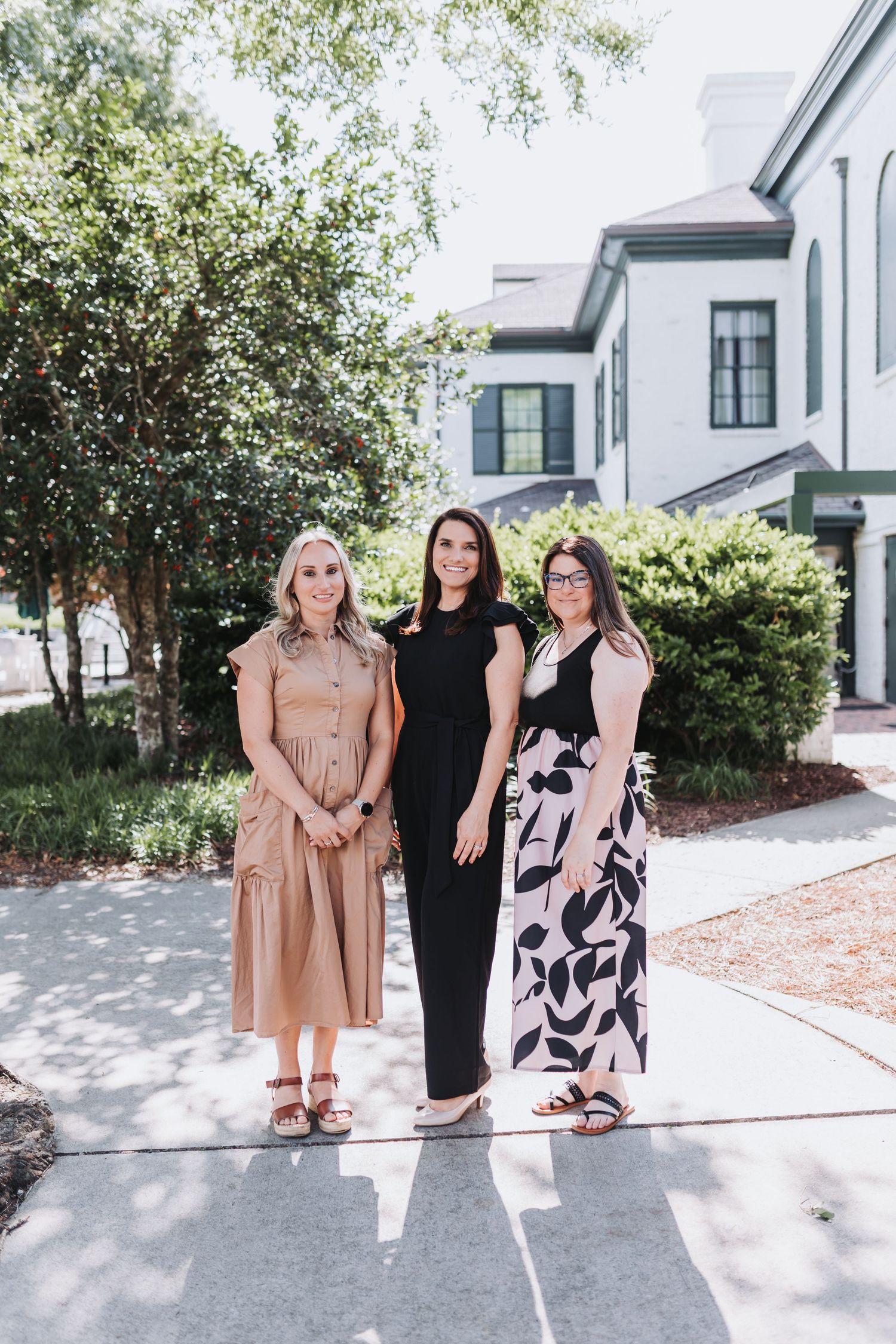 Three women smiling and posing together outdoors in front of a white building surrounded by lush green trees.