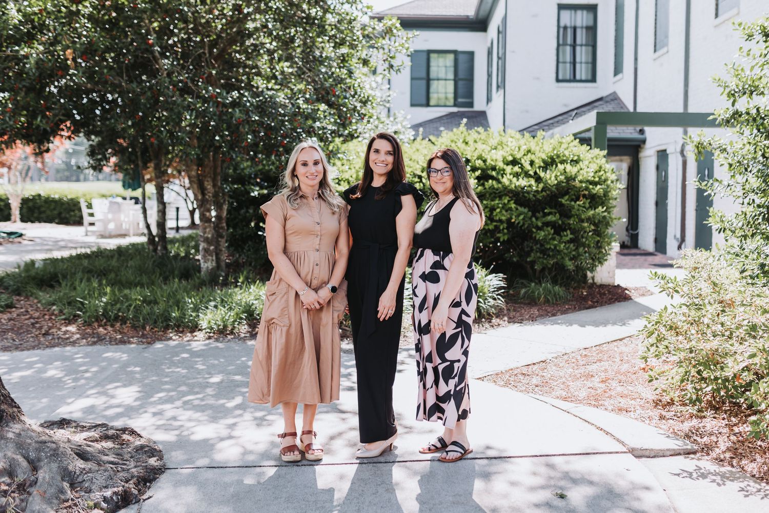 Three women posing together outdoors on a sunny day near a white building surrounded by green trees.