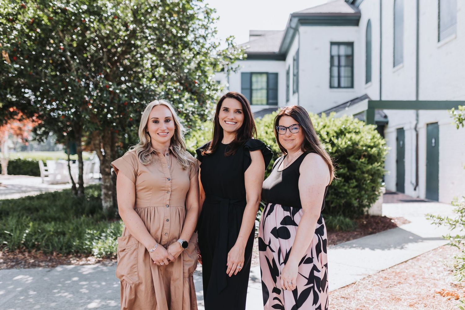 Three women in elegant dresses smile together outdoors near a white building surrounded by greenery.