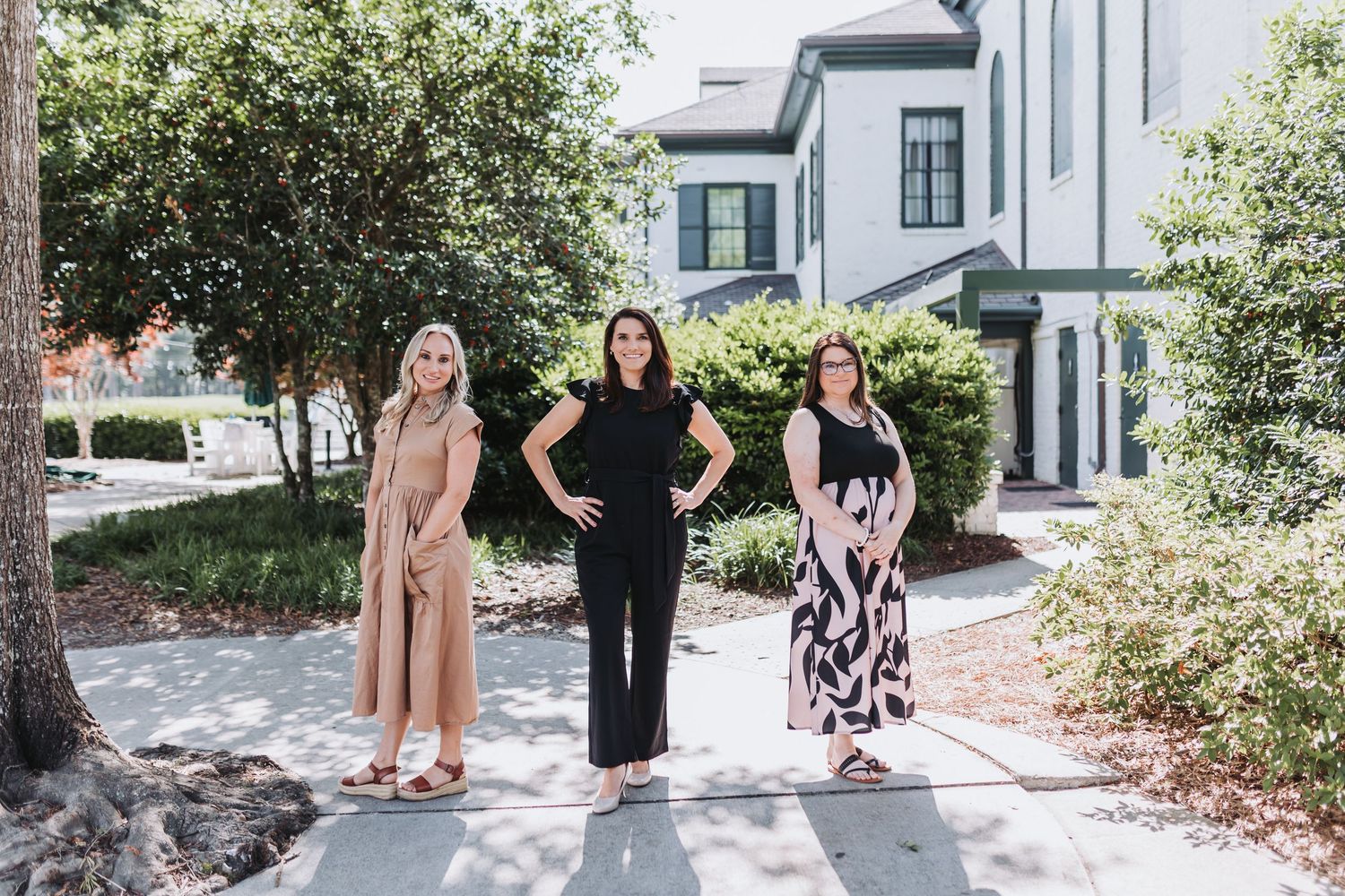 Three women pose outdoors on a sidewalk in front of a modern townhouse surrounded by lush green trees.