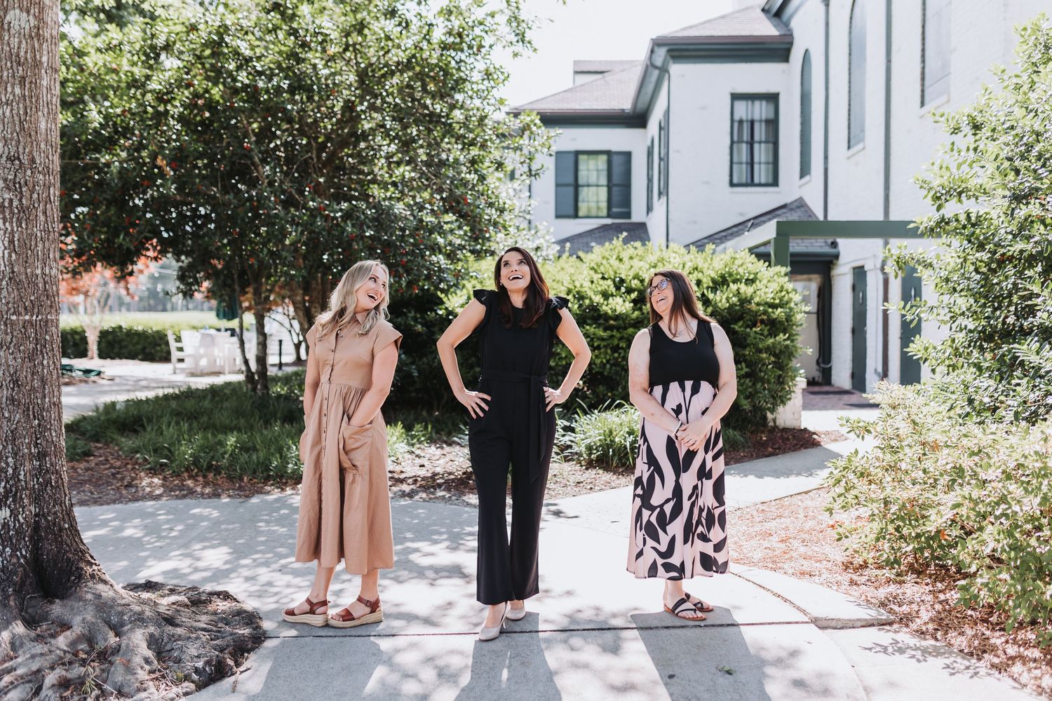 Three women in stylish outfits posing on a sunny sidewalk in front of a white house with lush greenery.