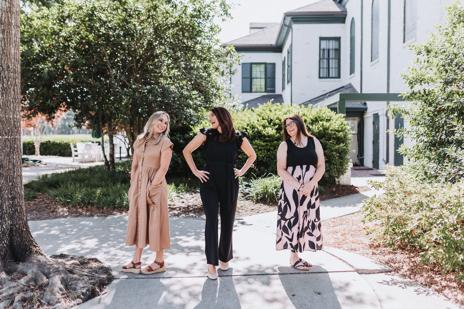 Three women standing outdoors on a sunny day near a white building surrounded by green shrubs and trees.