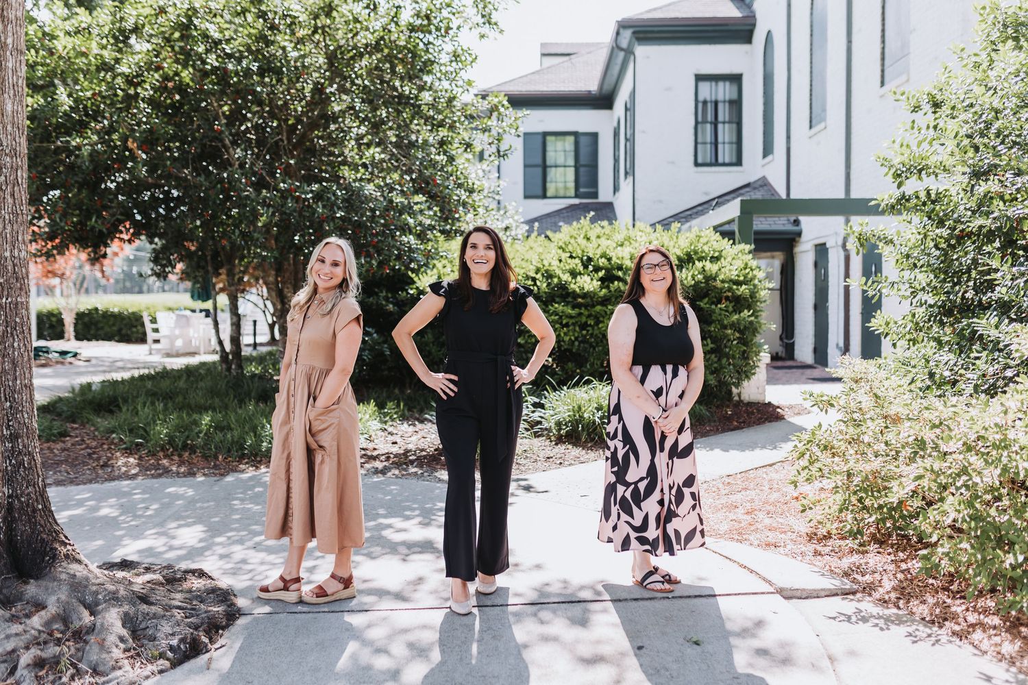 Three women pose confidently on a sunny walkway outside modern townhomes surrounded by lush green trees.