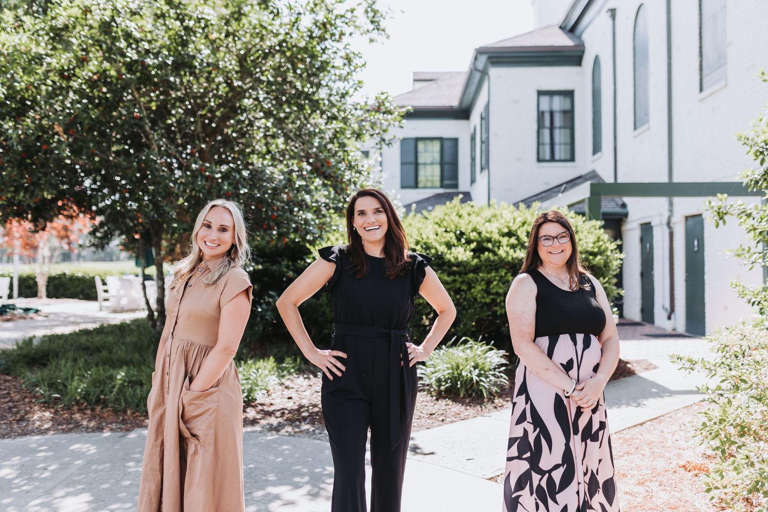 Three women smiling outdoors in front of a white building with lush green trees on a sunny day.