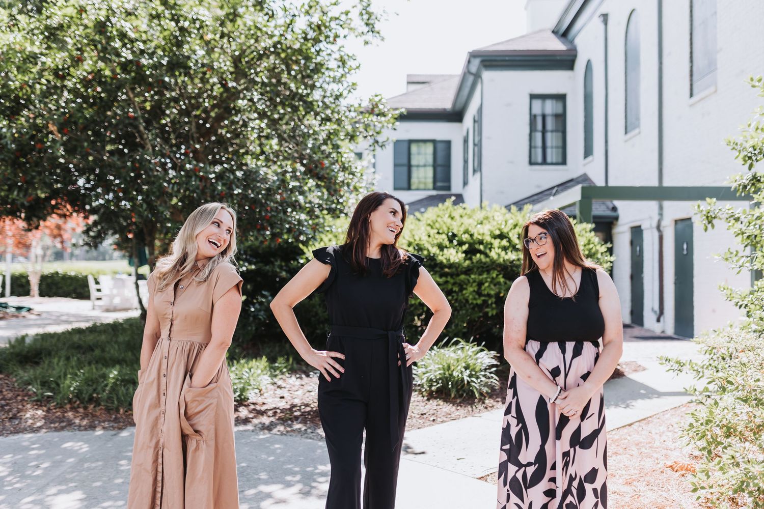Three women pose outdoors near a white building surrounded by greenery on a sunny day.