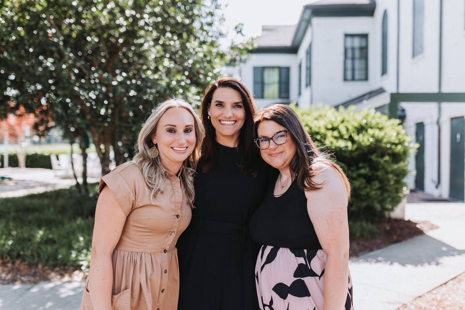 Three smiling women posing together outdoors near a building with greenery in the background.