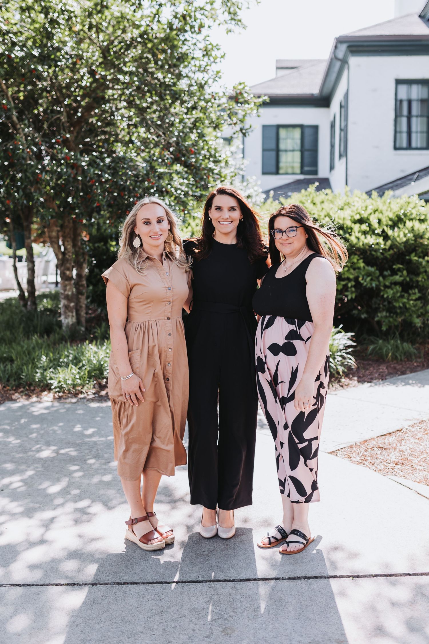 Three women smiling and posing together outdoors near trees and a building on a sunny day.