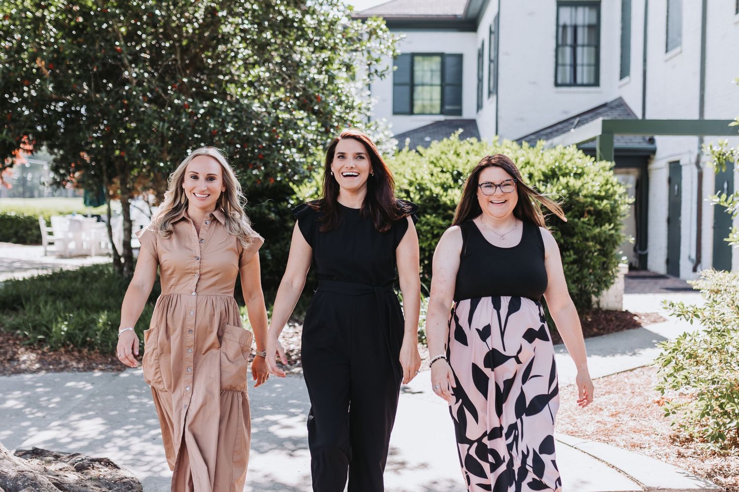 Three women walking together outdoors on a sunny day, smiling and dressed in stylish outfits near a white building.