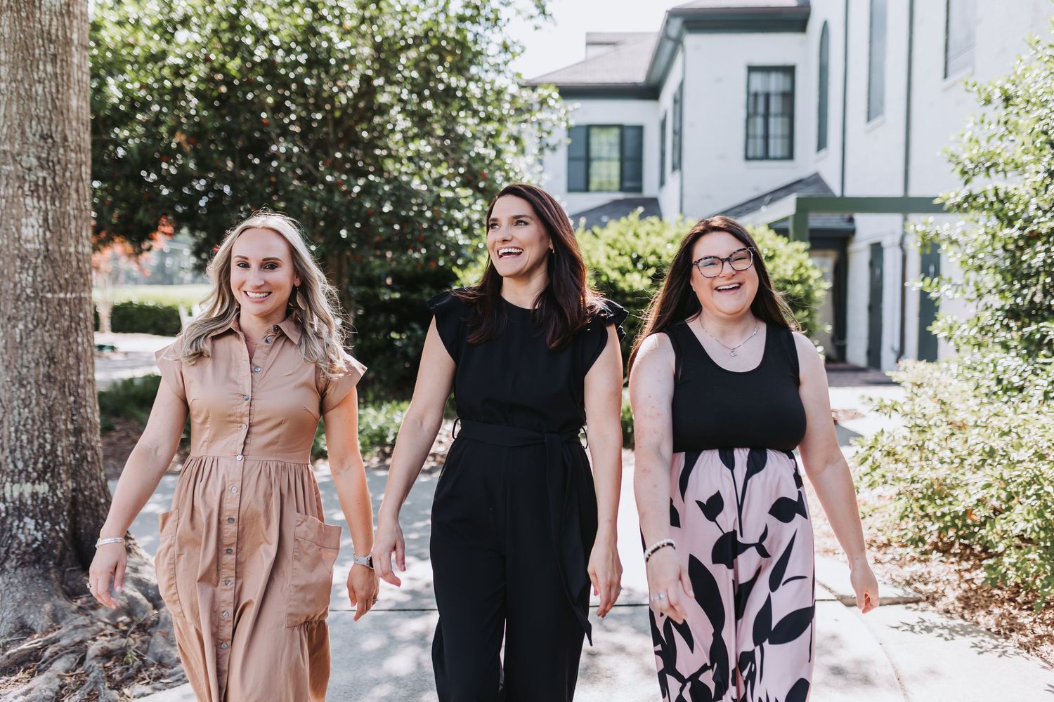 Three women walking together outdoors near a building, wearing stylish outfits and smiling on a sunny day.