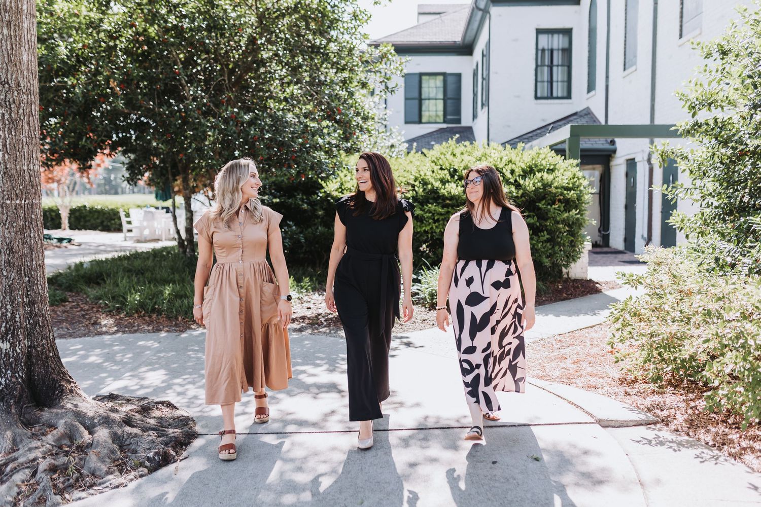 Three women walking together outdoors on a sunny day, wearing stylish summer outfits in neutral tones.