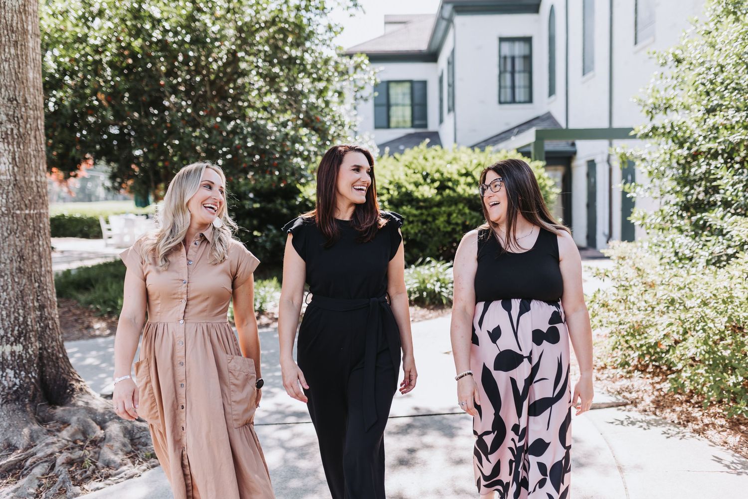 Three stylish women in elegant dresses walking together outdoors on a sunny day near a white building.