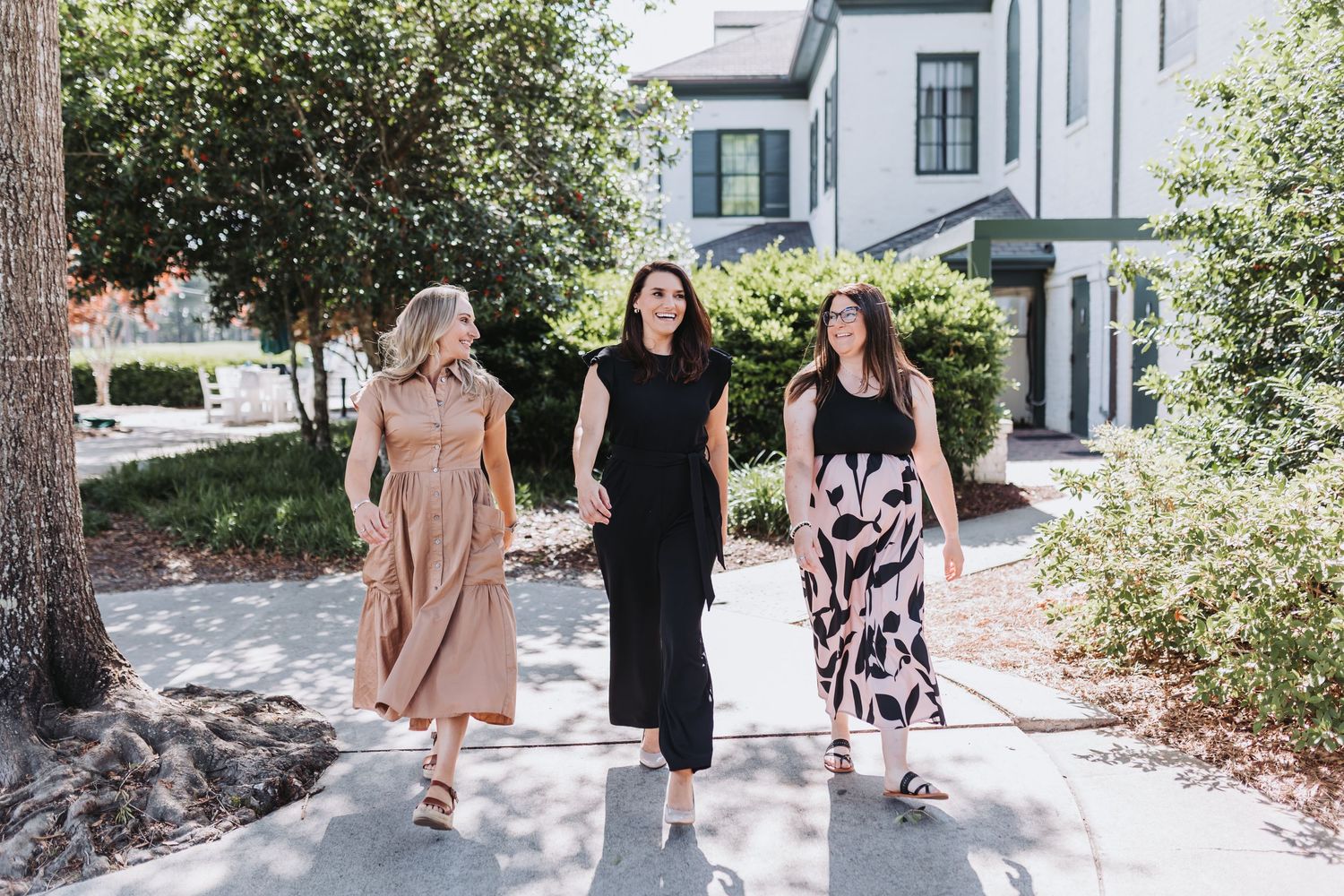 Three stylish women walking together outdoors on a sunny day, wearing elegant dresses in black, beige, and floral print.