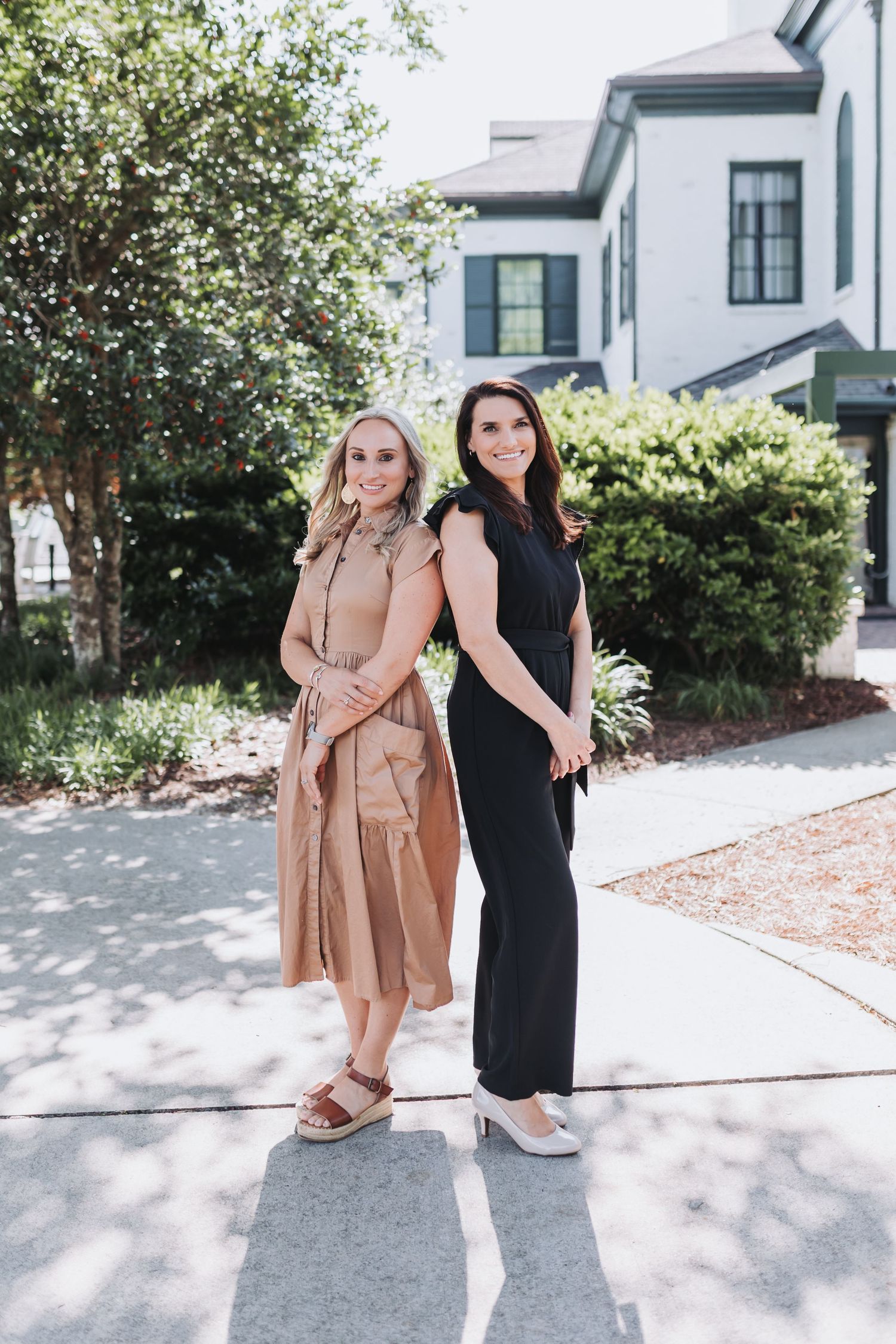 Two women posing outdoors, one in a tan dress and one in a black jumpsuit, standing in front of a white building.