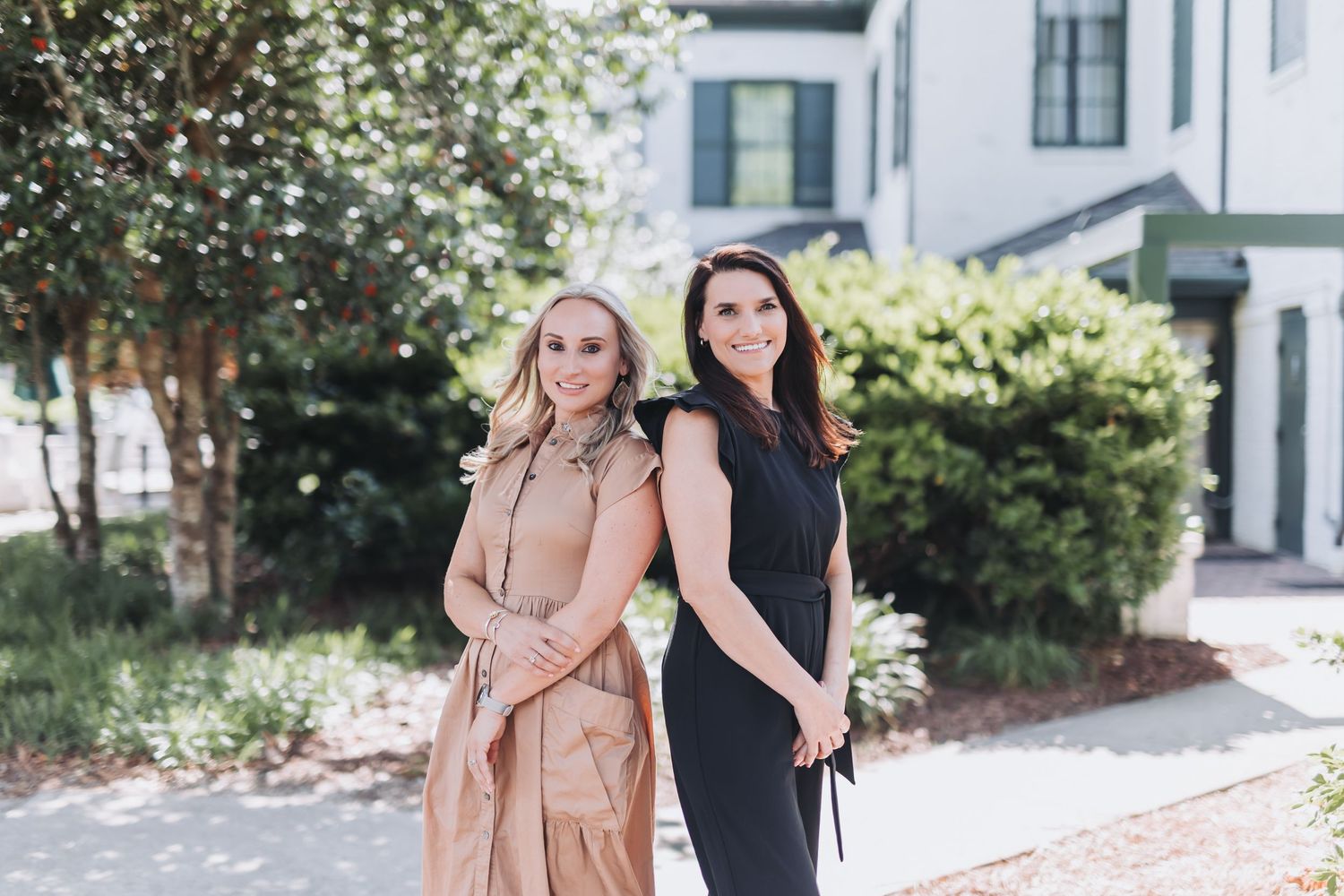 Two women posing back-to-back outdoors near a white building, one in a pink dress and one in a black dress.