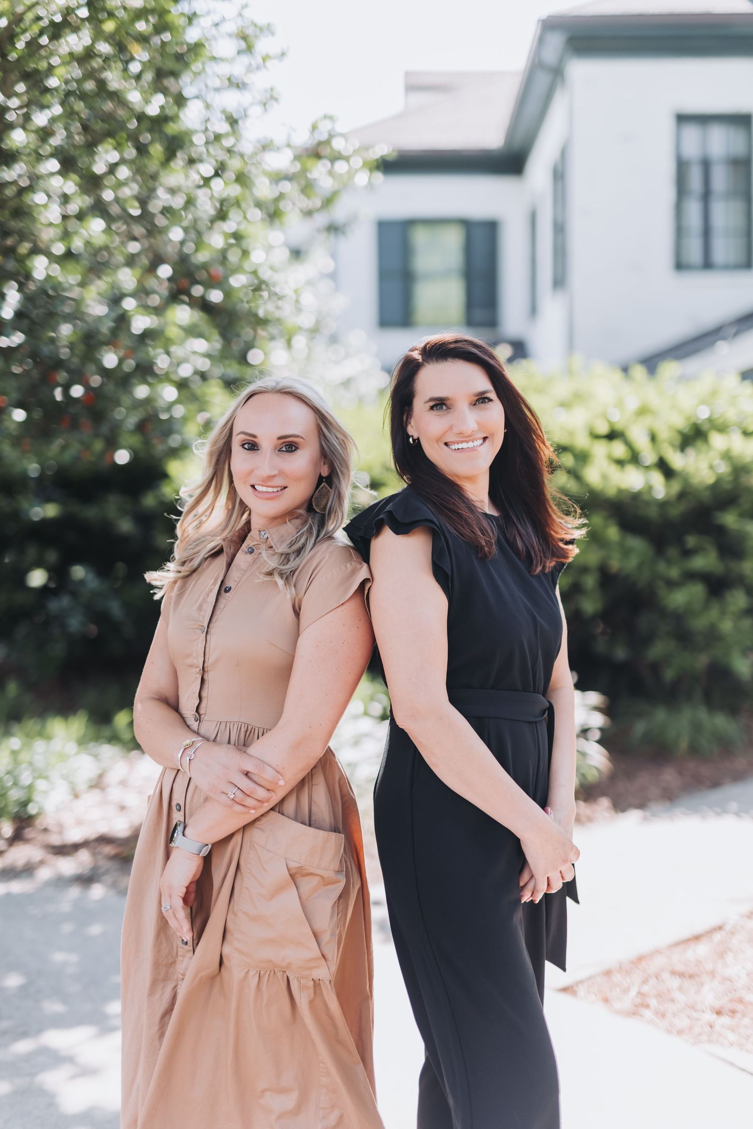 Two smiling women in elegant dresses pose back-to-back outdoors near lush greenery and a white building.