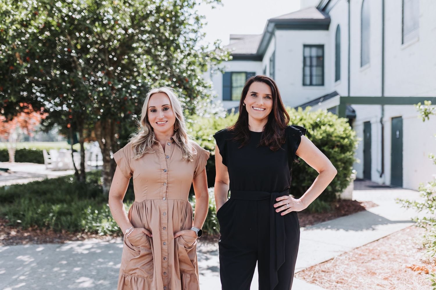 Two professional women smiling outdoors near a white building, one in a tan dress and one in a black outfit.
