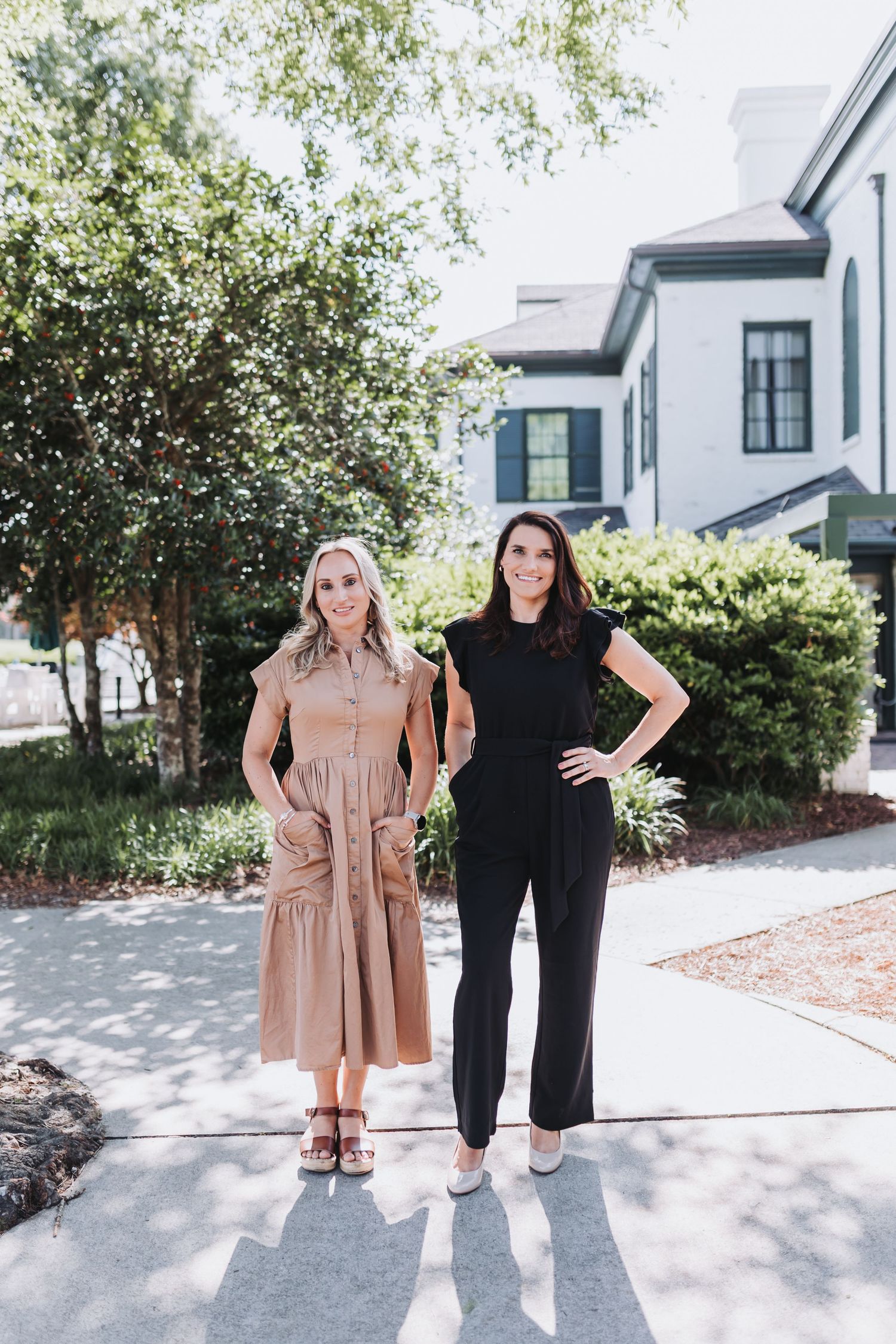 Two women smiling outdoors, one in a tan dress and one in a black jumpsuit, standing on a sidewalk near a house.