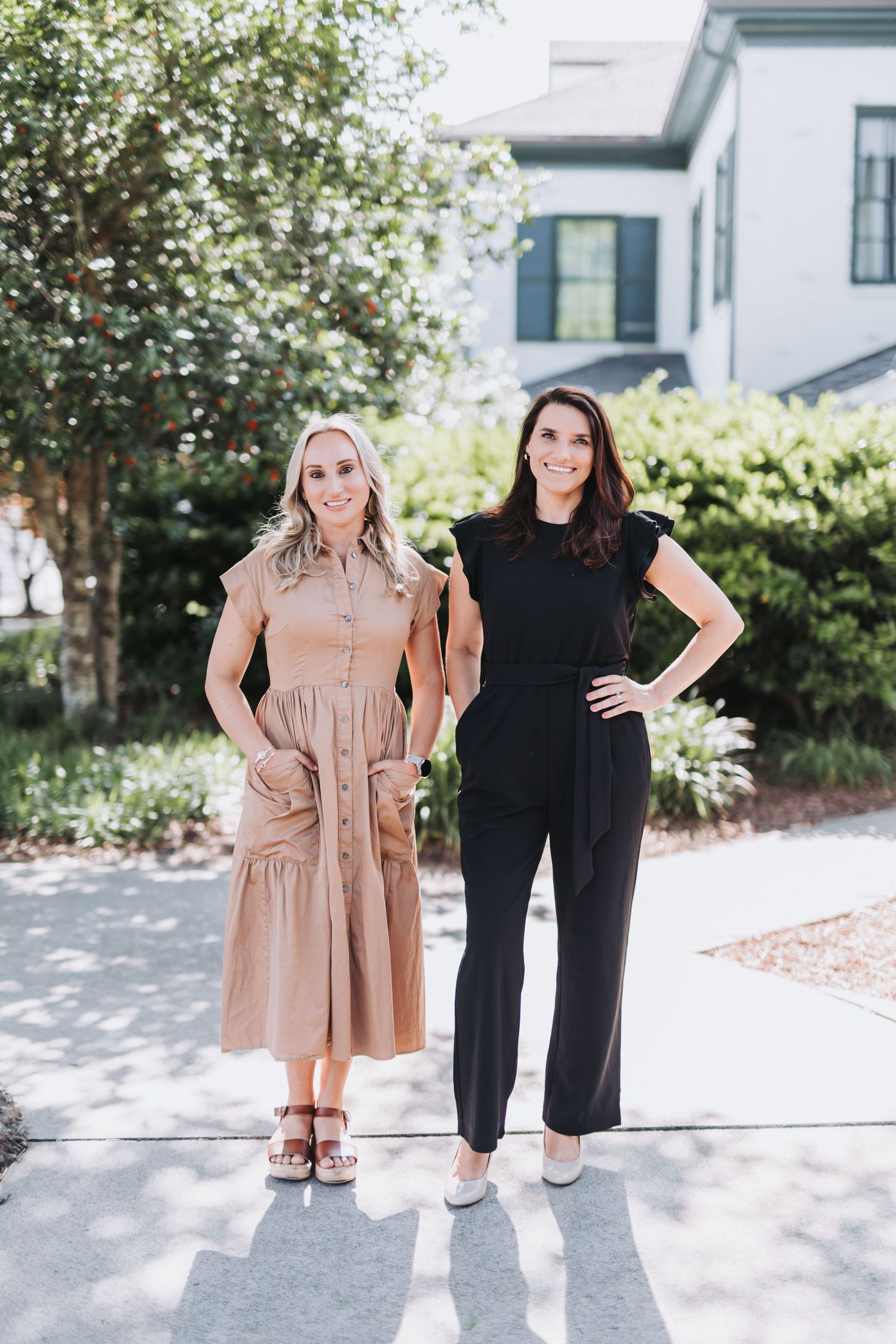 Two women smiling outdoors, one in a tan midi dress and one in a black jumpsuit, standing on a sunny sidewalk.
