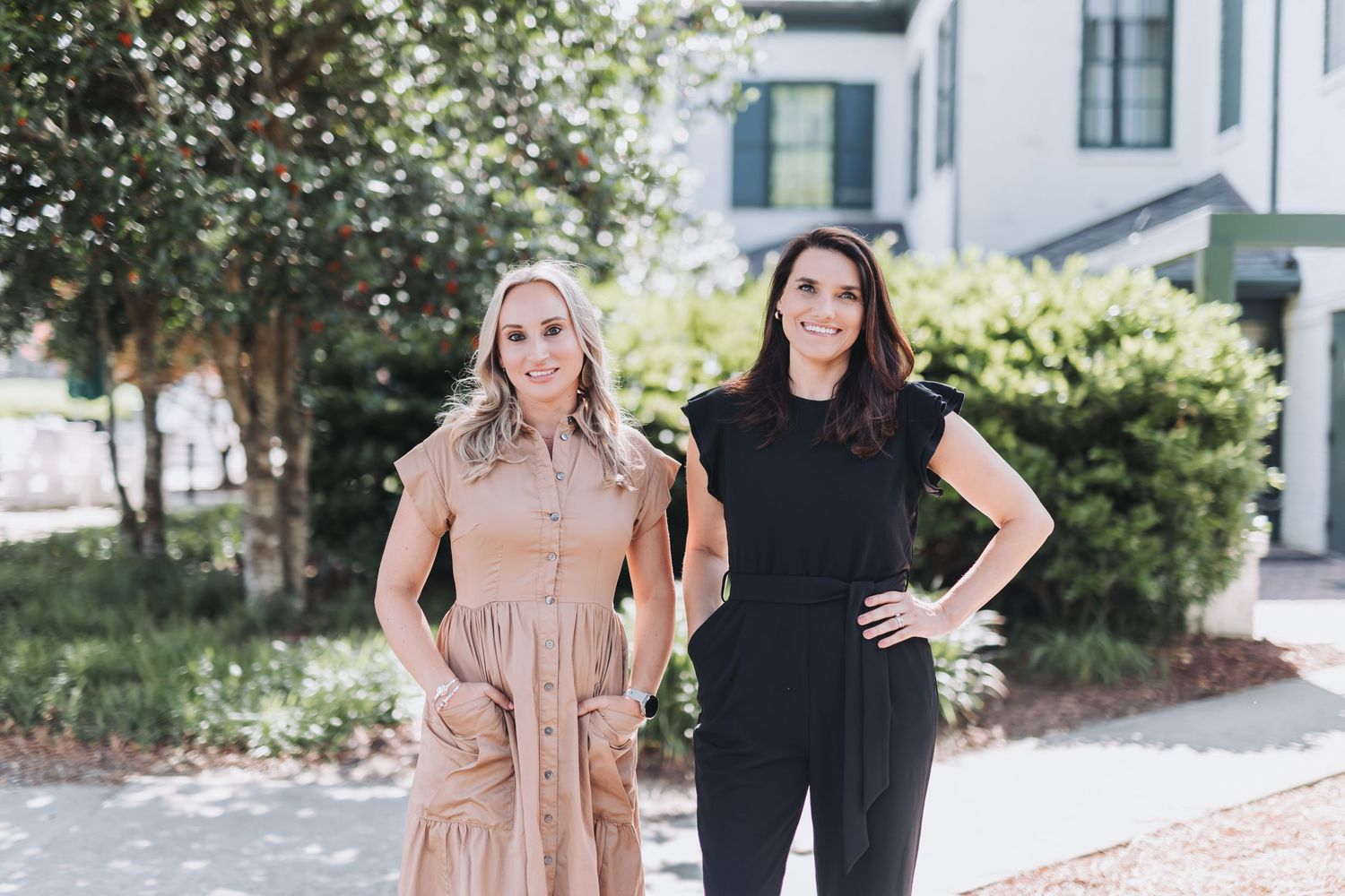 Two women smiling outdoors in front of a white building, one in a tan dress and one in a black outfit.