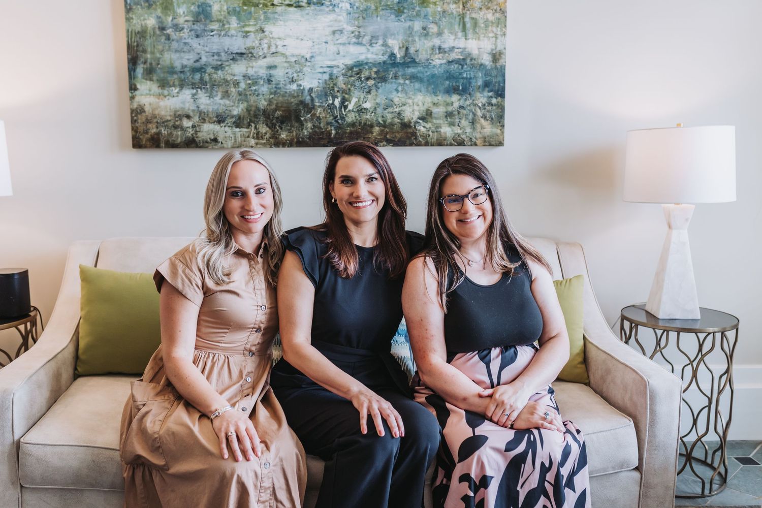 Three women smiling together on a white sofa in an elegant living room with abstract wall art.