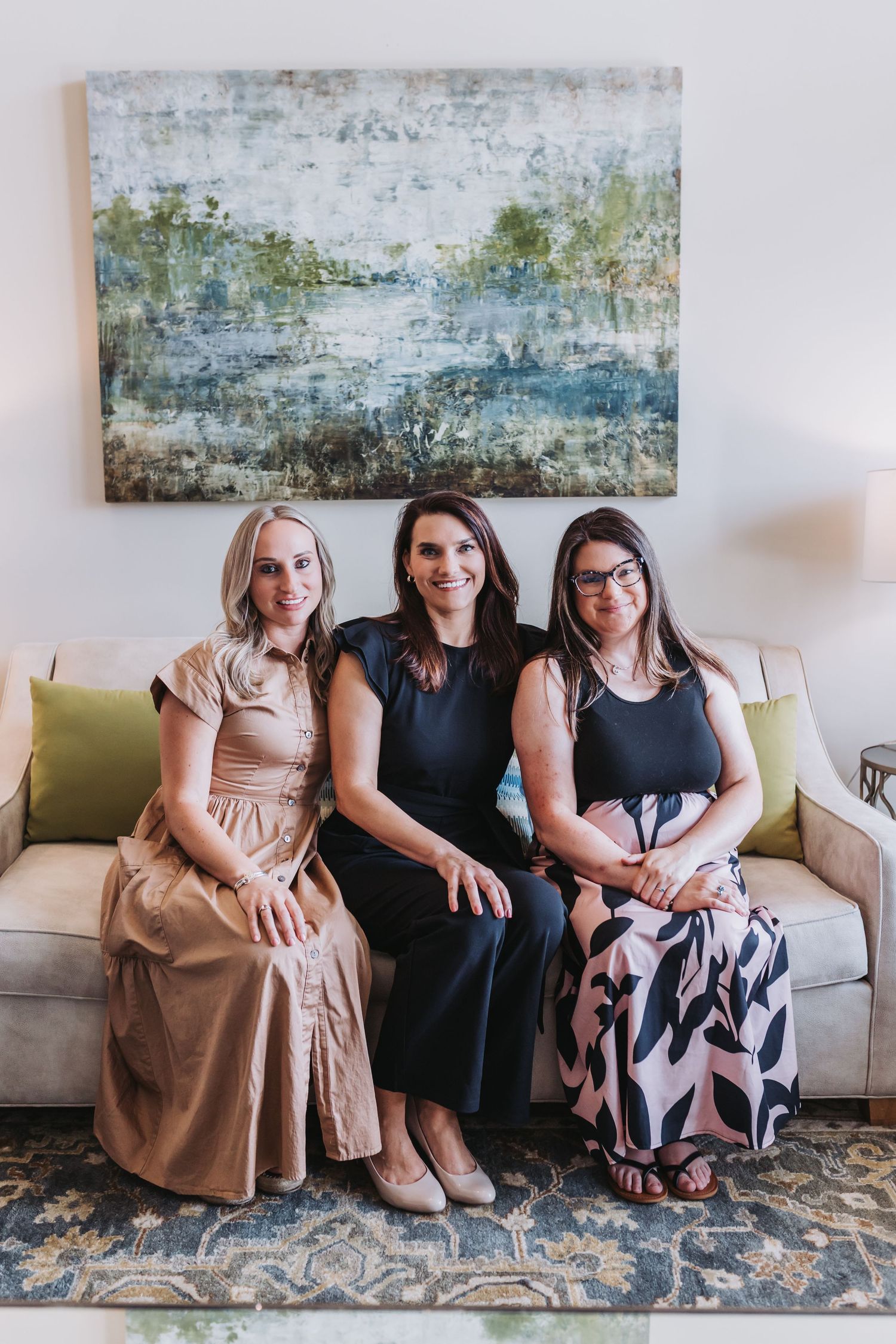 Three women sitting together smiling on a beige sofa with abstract landscape painting on wall behind them.