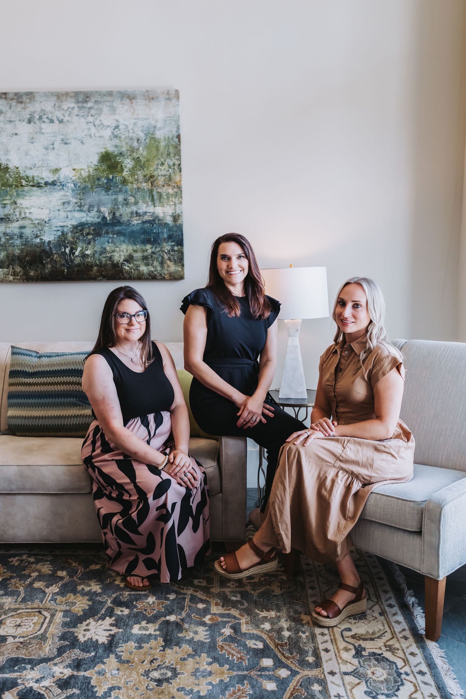 Three women smiling and posing together on modern sofas in a bright, elegantly decorated living room space.