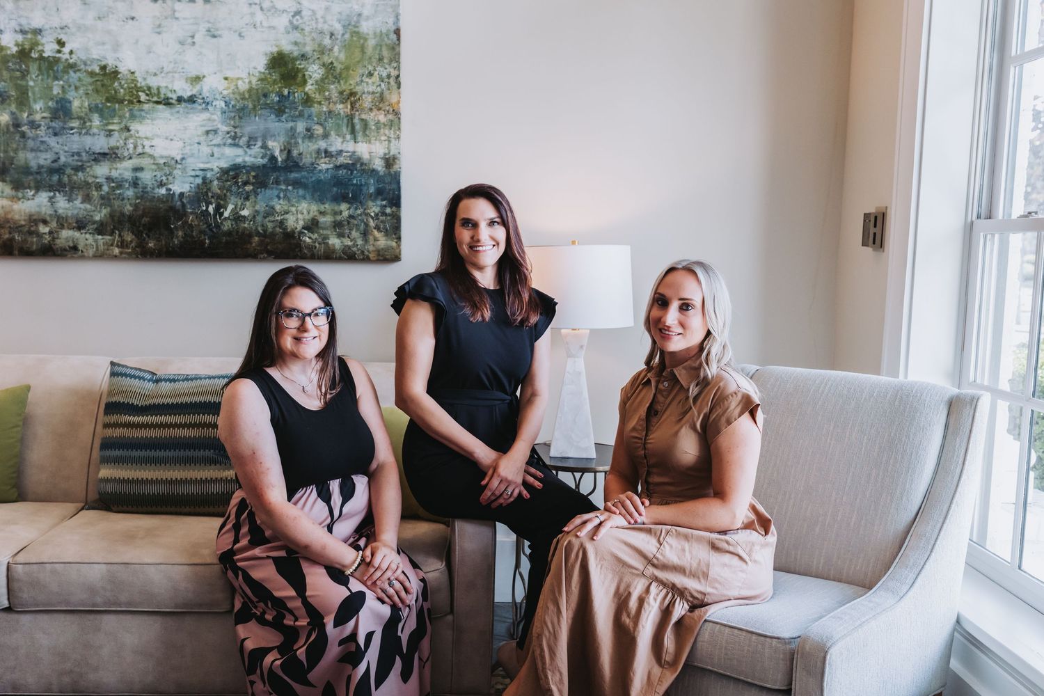 Three professional women sitting together on a beige sofa in an elegant, well-lit office or living room setting.