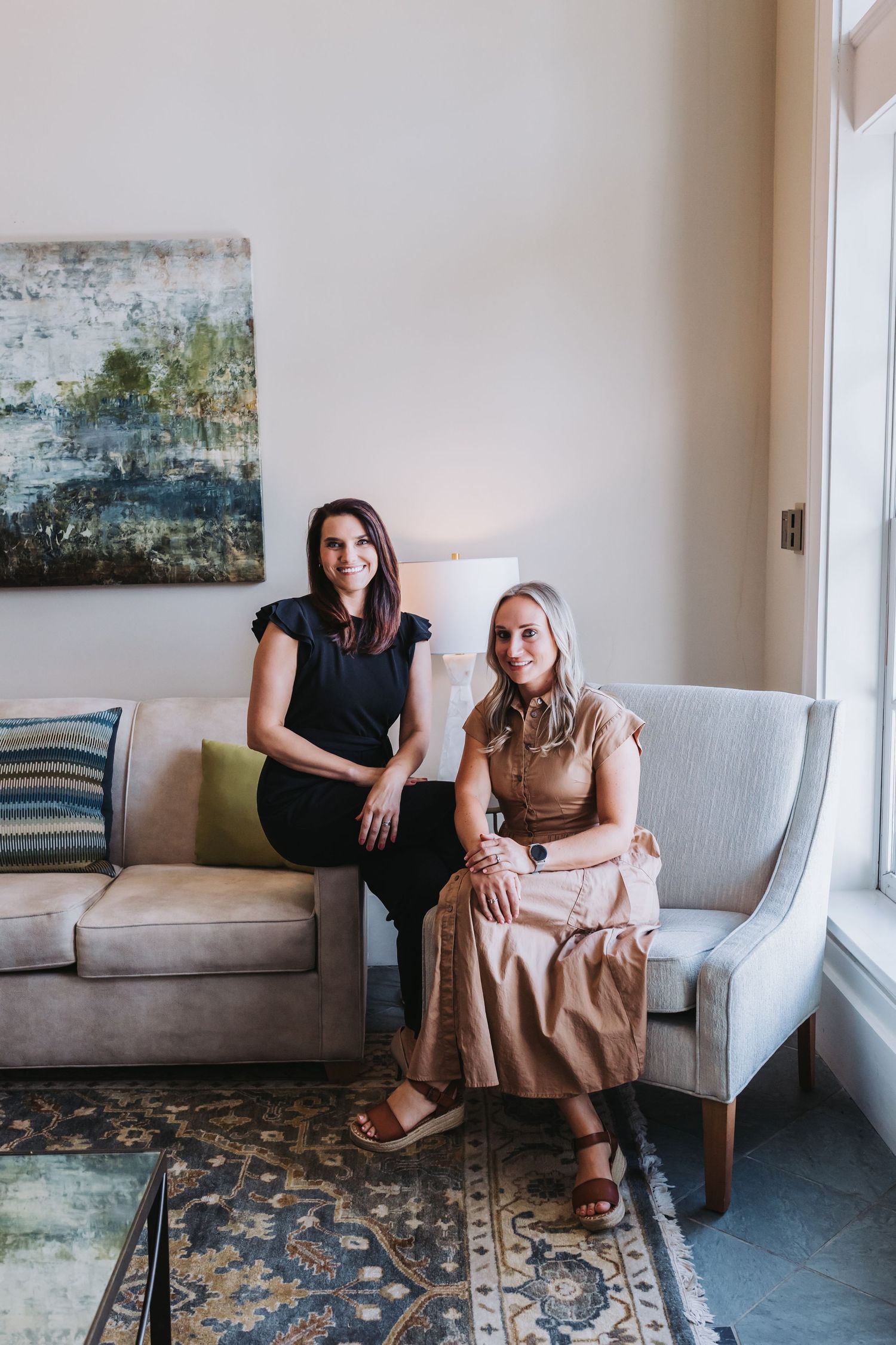 Two women sitting and smiling in a elegantly decorated living room with abstract art and neutral toned furniture.