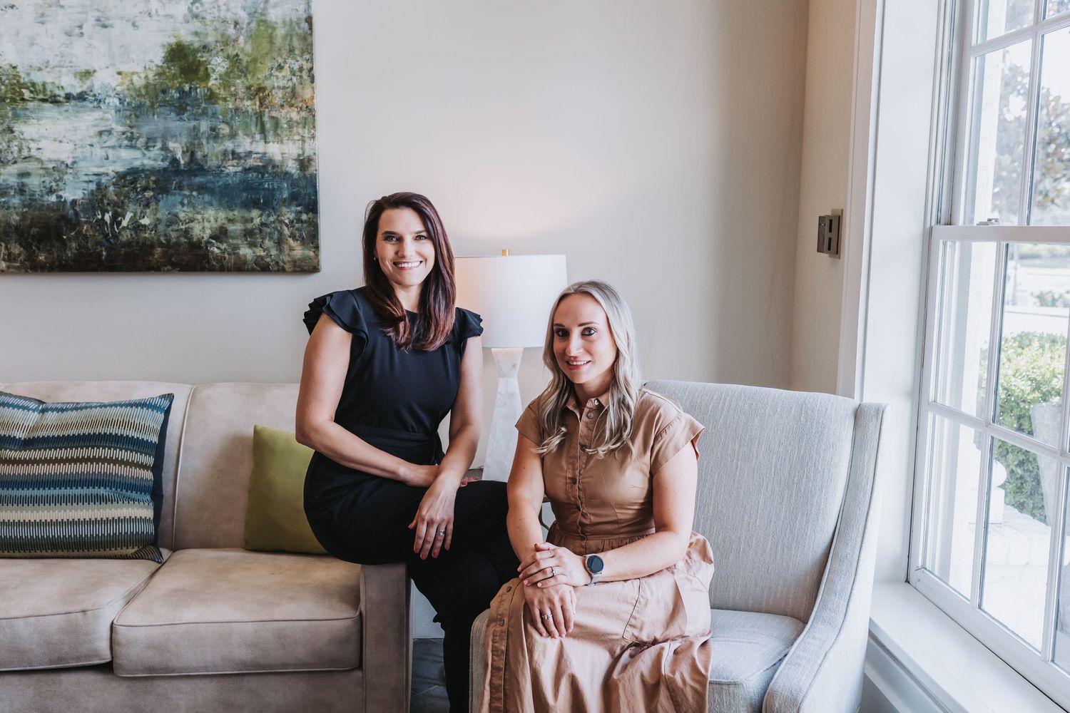 Two professional women smiling and posing together in a bright, elegantly decorated living room.