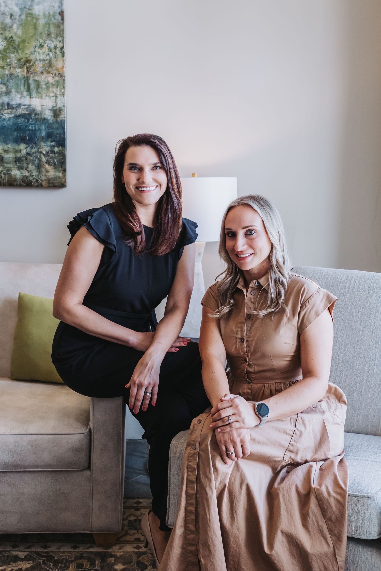 Two smiling women sitting on a light gray sofa in a modern interior, one in black and one in beige dress.