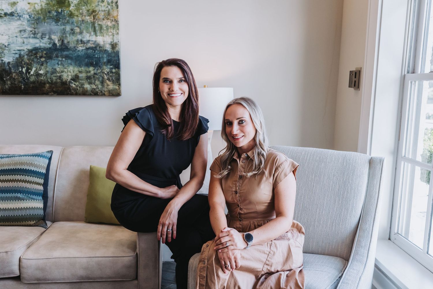Two professional women sitting together on a light gray sofa in a modern, elegantly decorated living room.