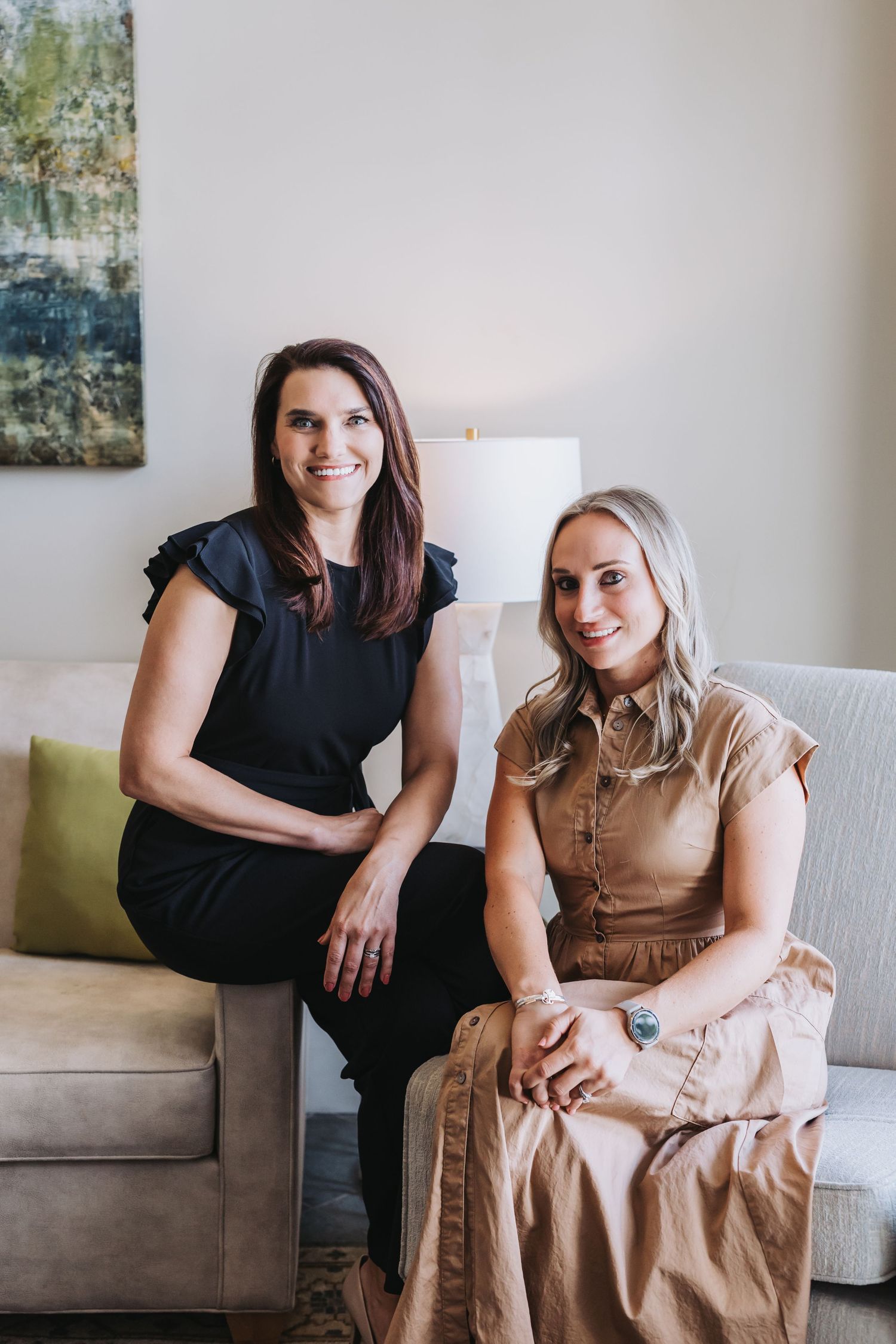 Two women smiling and sitting together on a couch in a modern, well-lit interior setting.