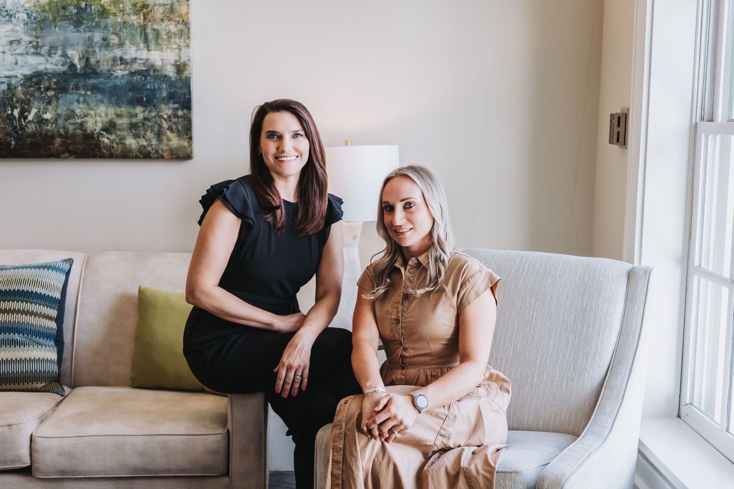 Two women smiling and sitting together on a light gray sofa in a bright, elegantly decorated living room.