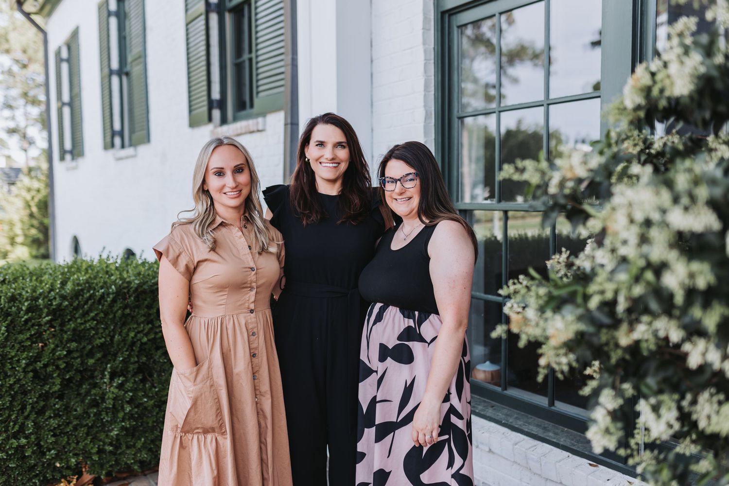 Three women smiling and posing together outdoors in front of a white building with lush greenery.