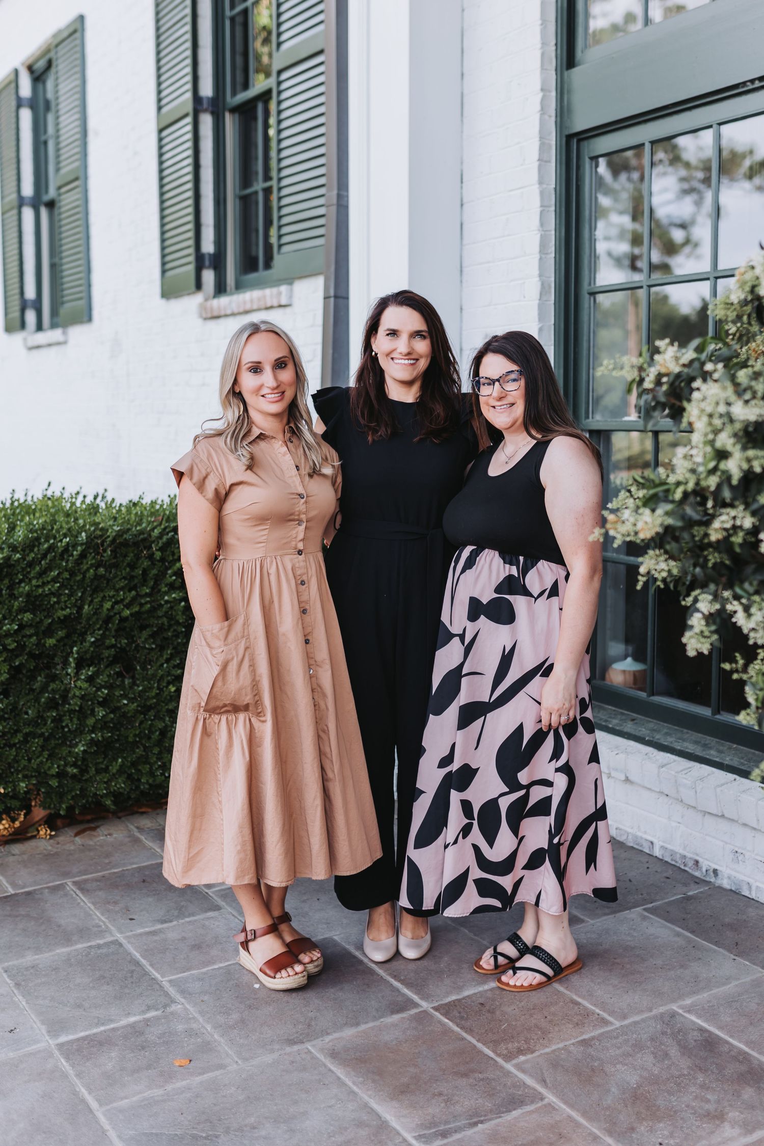 Three women in stylish dresses smiling together outside a white brick building with green shutters.