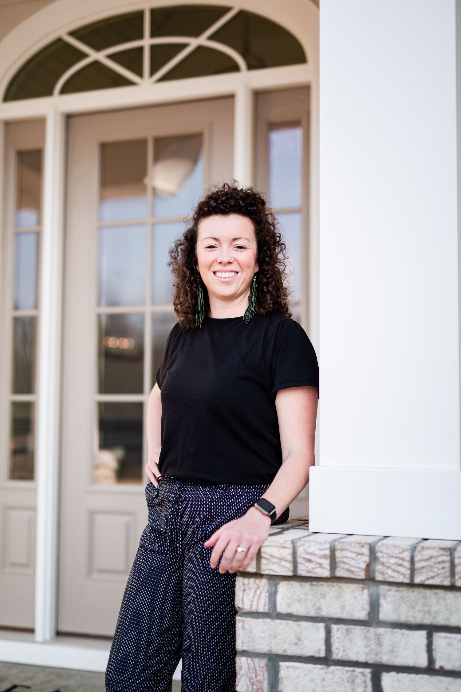 Woman with curly hair in black top and polka dot pants smiling outdoors near brick wall and white door.