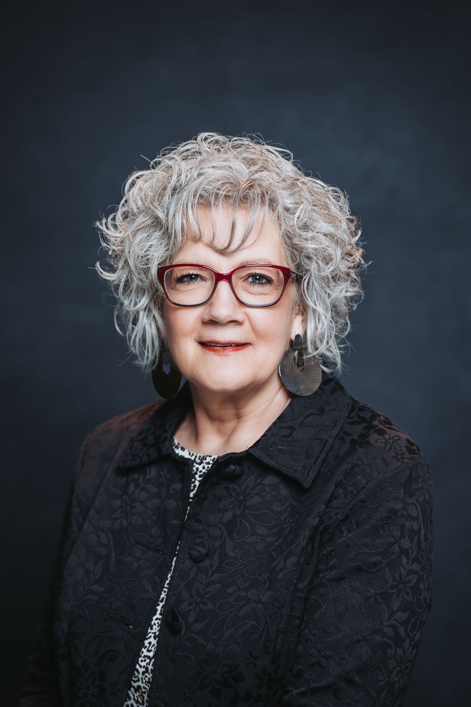 Older woman with curly gray hair, red glasses, and dark floral jacket smiling confidently against a dark background.