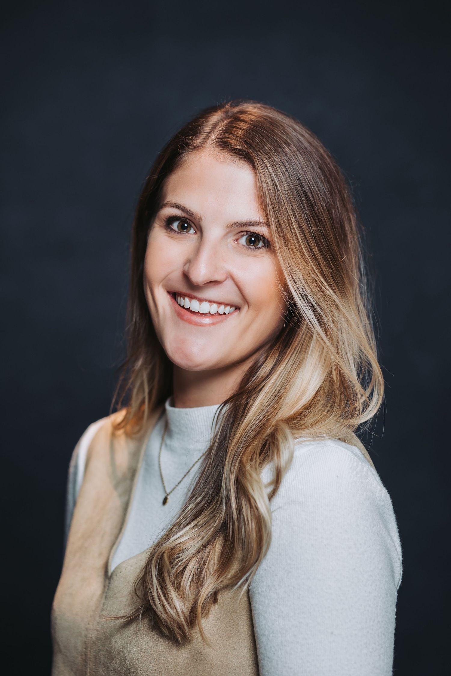 Smiling woman with long brown hair wearing a light sweater and necklace against a dark background.