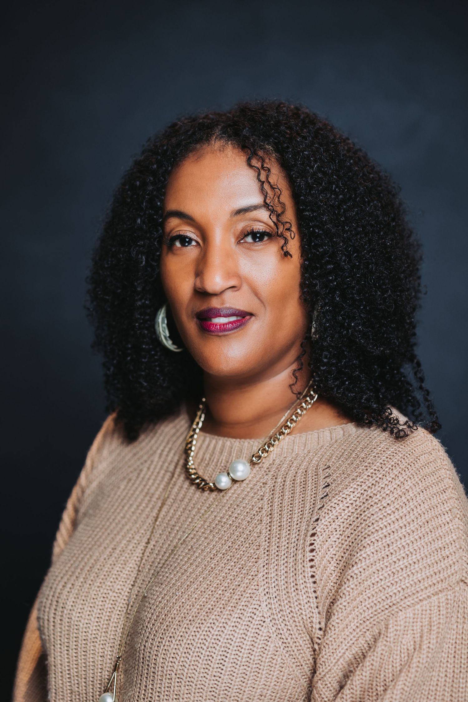 Professional headshot of a woman with curly hair wearing a beige sweater and gold necklace against a dark background.