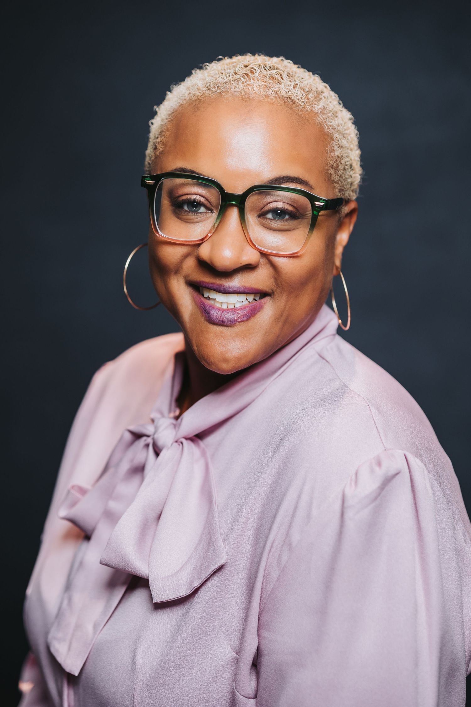 Smiling woman with short blonde hair, green glasses, hoop earrings, and a lavender bow blouse poses against a dark background.