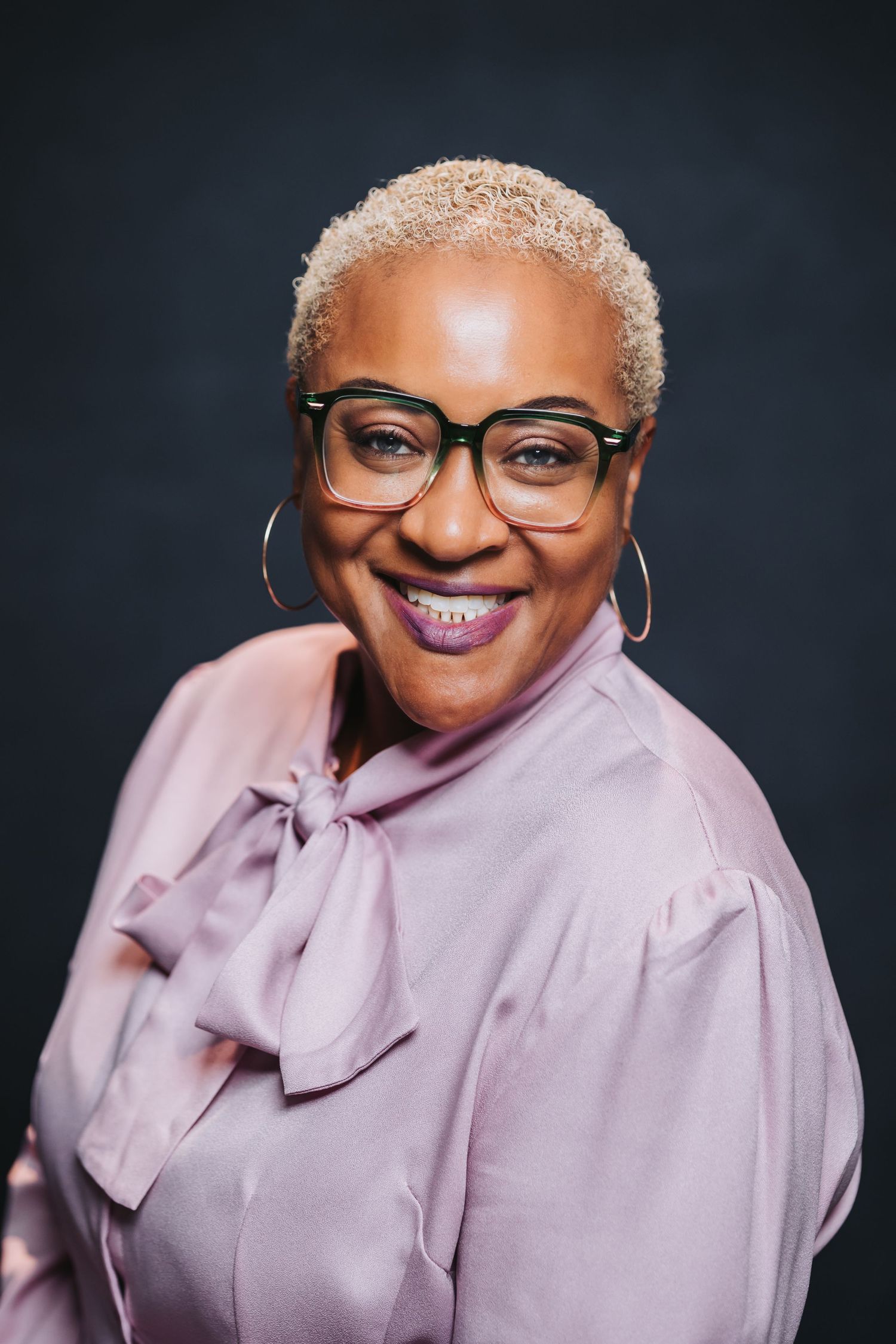 Smiling woman with short blonde hair, glasses, and hoop earrings wearing a pink blouse against dark background.