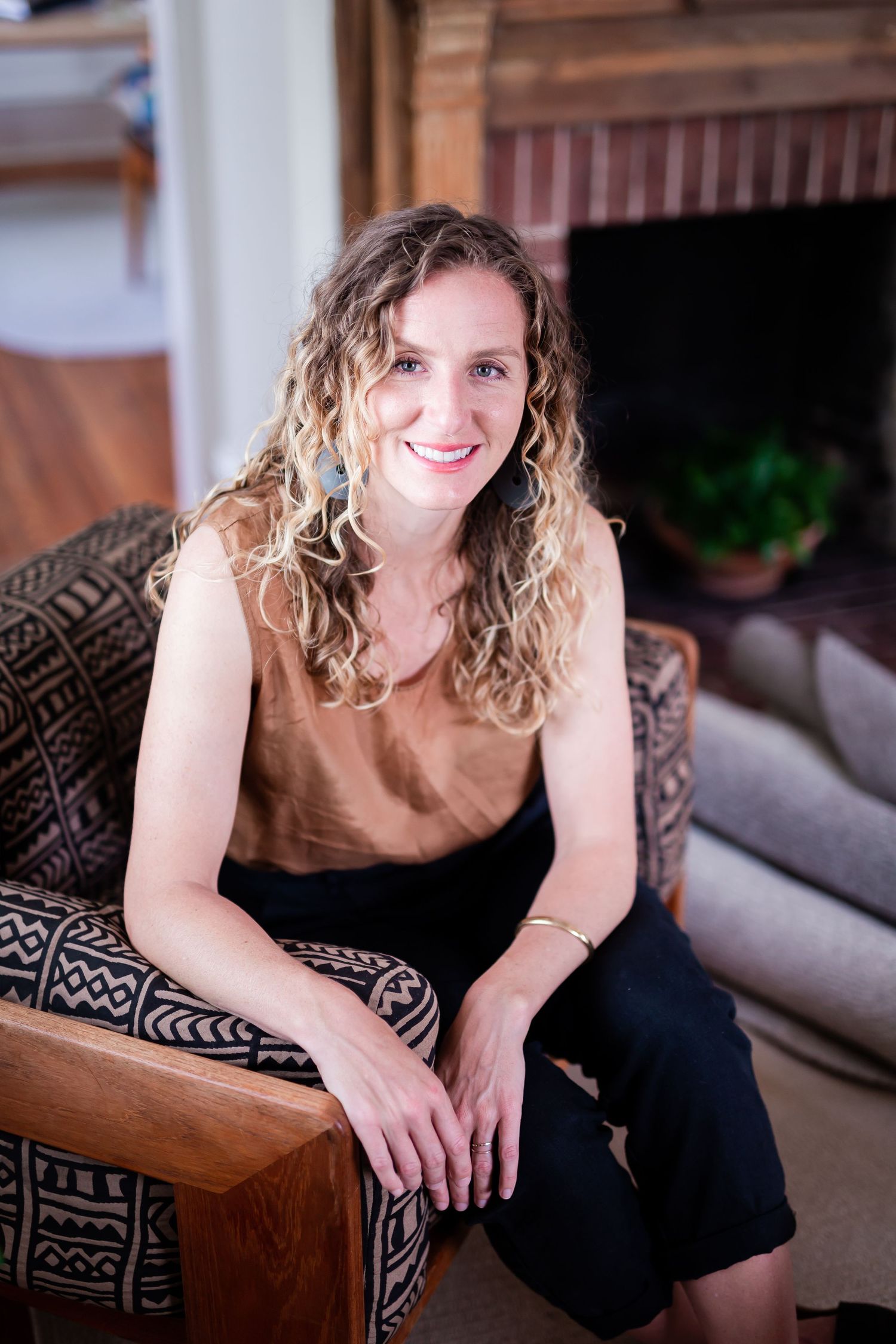 Smiling woman with curly blonde hair sitting in a patterned armchair near a fireplace, wearing a brown top and black pants.