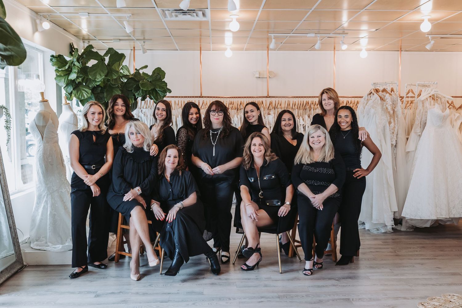 A team of women in black outfits posing together in an elegant bridal boutique surrounded by wedding gowns.
