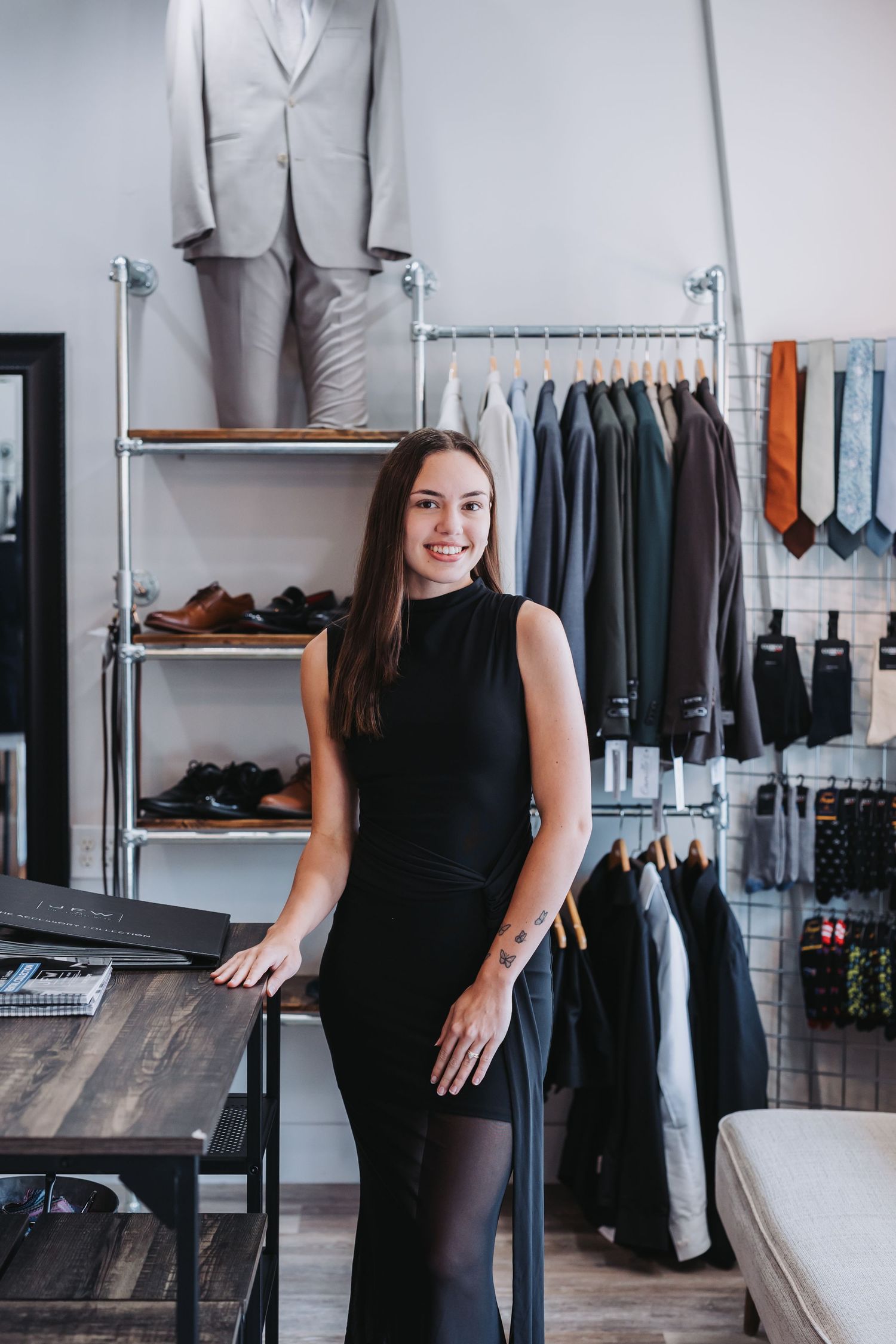 Woman in black dress smiling in a men's clothing boutique with suits, ties, and shoes on display.